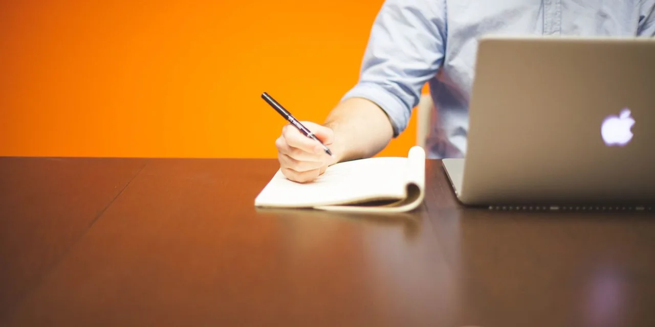Adult writing notes in a notebook beside a laptop on a wooden desk with orange background.
