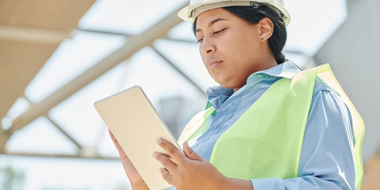 Confident female engineer in hard hat using a tablet computer at an outdoor construction site.