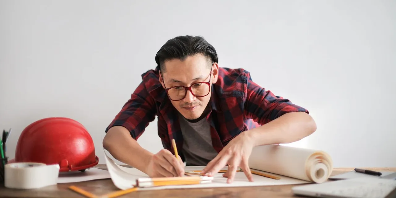 Inspired young male engineer in glasses drawing draft on paper bending over table against white wall